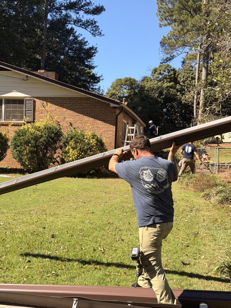 Installer carrying a brown gutter run across the front lawn of a brick home in Smyrna