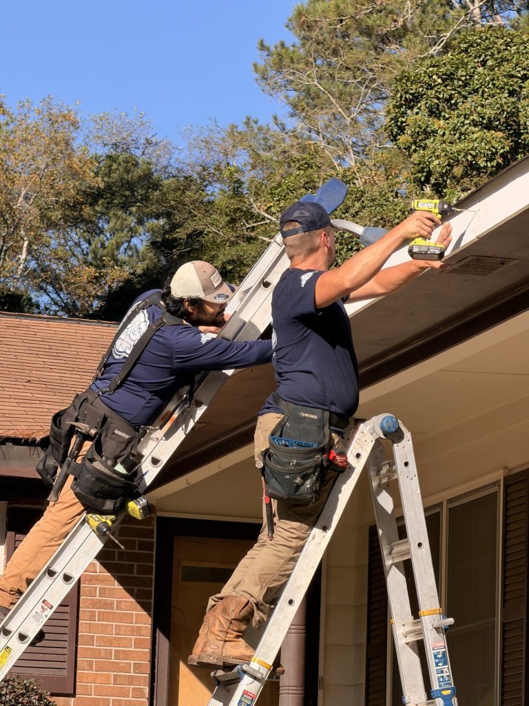 Installers fastening a new brown gutter along the roofline of a brick home in Smyrna