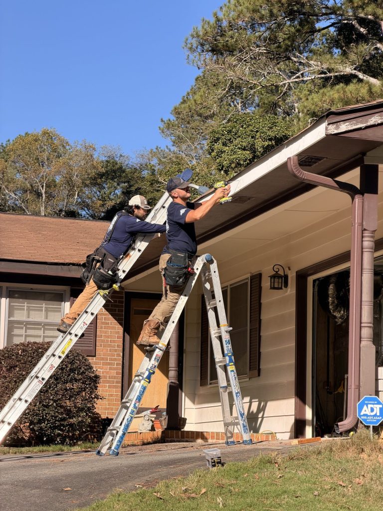 Two installers on ladders hanging a brown seamless gutter on a brick ranch home in Smyrna